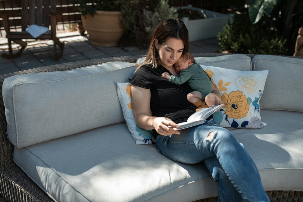 A serene moment of a mother reading while holding her sleeping baby on an outdoor sofa.