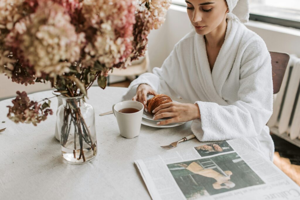 A woman in a bathrobe having breakfast indoors with a croissant and tea.