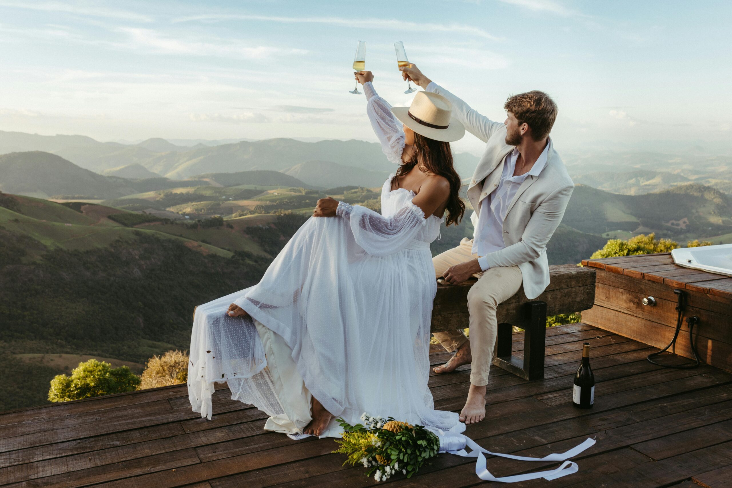 A couple in wedding attire toasts to their new life on a scenic mountain deck.