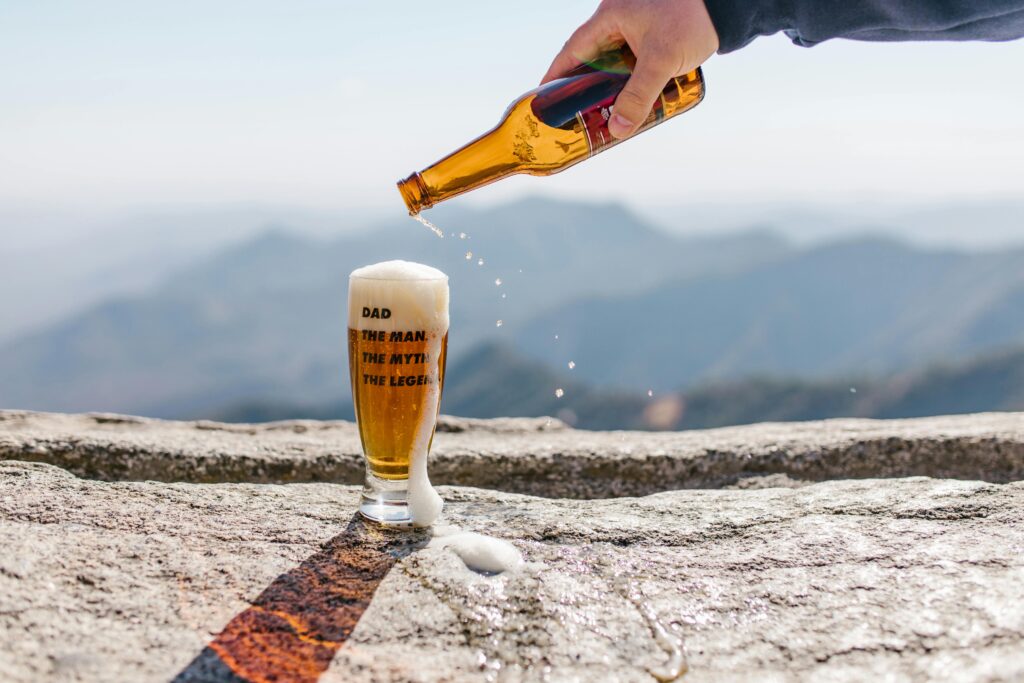 Hand pouring beer into glass on a mountain with a scenic view and Father's Day message.