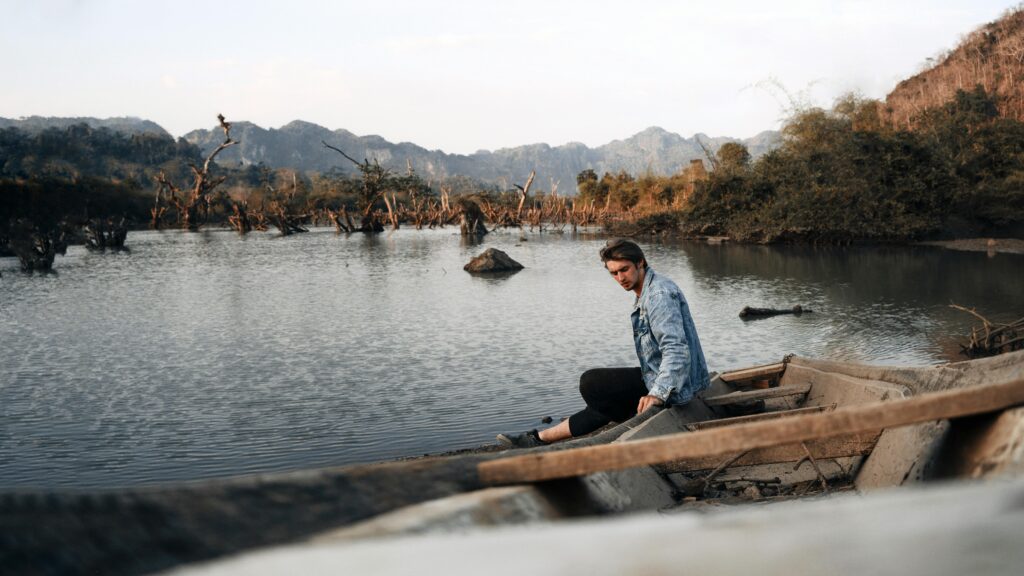 A man in a denim jacket sits in a boat on a serene Laos river with a mountainous backdrop.