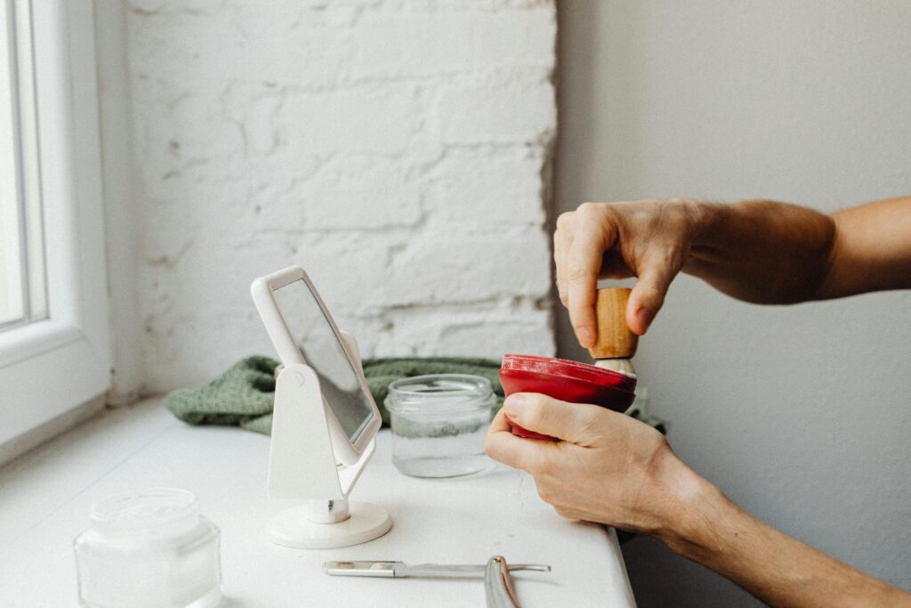 A man's hands prepare shaving cream using a brush by a window.