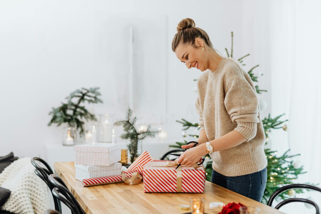 A woman joyfully wraps Christmas presents in a festive, cozy indoor setting.