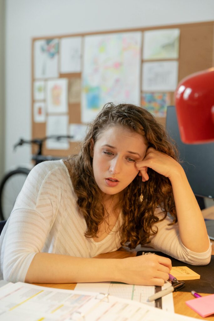 A tired woman leans on a desk, looking stressed in an office setting with papers.