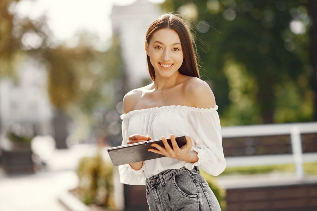 A cheerful woman holding a tablet in a sunny park, exuding happiness and casual style.
